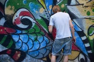 foto en el proceso de dibujar un patrón de graffiti en un antiguo muro de hormigón. un joven rubio de pelo largo dibuja un dibujo abstracto de diferentes colores. concepto de arte callejero y vandalismo