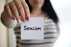 Young sad girl shows a white sticker. Caucasian brunette holding a sheet of paper with message. Sexism photo