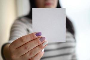 Young sad girl shows a white sticker. Caucasian brunette holding a blank sheet of paper as a template for your text photo