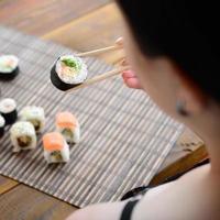 Brunette girl with chopsticks holds a sushi roll on a bamboo straw serwing mat background. Traditional Asian food photo