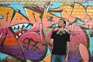 Young graffiti artist with gas mask on his neck throw his spray can against colorful pink graffiti on brick wall. Street art and contemporary painting process photo