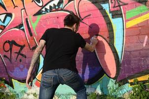 Young graffiti artist with backpack and gas mask on his neck paints colorful graffiti in pink tones on brick wall. Street art and contemporary painting process photo