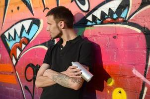 joven artista de graffiti caucásico en camiseta negra con lata de aerosol plateado cerca de graffiti colorido en tonos rosas en la pared de ladrillo. proceso de arte callejero y pintura contemporánea foto