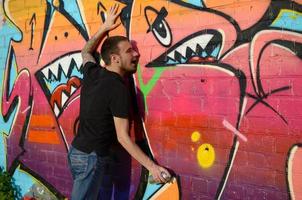 Young caucasian graffiti artist in black t-shirt with silver aerosol spray can near colorful graffiti in pink tones on brick wall. Street art and contemporary painting process photo