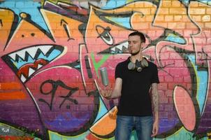 Young graffiti artist with gas mask on his neck throw his spray can against colorful pink graffiti on brick wall. Street art and contemporary painting process photo