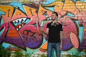 el joven artista de graffiti con máscara de gas en el cuello lanza su lata de aerosol contra el colorido graffiti rosa en la pared de ladrillo. proceso de arte callejero y pintura contemporánea foto