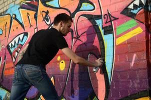 Young graffiti artist with backpack and gas mask on his neck paints colorful graffiti in pink tones on brick wall. Street art and contemporary painting process photo