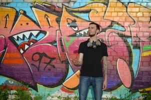 el joven artista de graffiti con máscara de gas en el cuello lanza su lata de aerosol contra el colorido graffiti rosa en la pared de ladrillo. proceso de arte callejero y pintura contemporánea foto