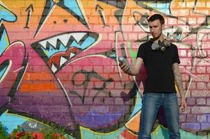 Young graffiti artist with gas mask on his neck throw his spray can against colorful pink graffiti on brick wall. Street art and contemporary painting process photo