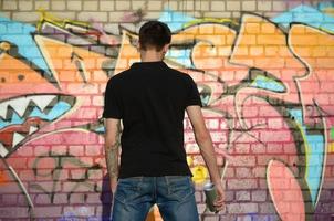 Young graffiti artist with backpack and gas mask on his neck paints colorful graffiti in pink tones on brick wall. Street art and contemporary painting process photo