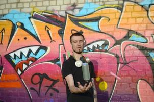 Young graffiti artist with gas mask on his neck throw his spray can against colorful pink graffiti on brick wall. Street art and contemporary painting process photo