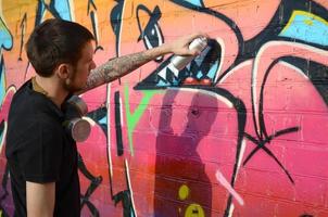 Young graffiti artist with backpack and gas mask on his neck paints colorful graffiti in pink tones on brick wall. Street art and contemporary painting process photo