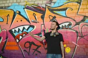 Young graffiti artist with gas mask on his neck throw his spray can against colorful pink graffiti on brick wall. Street art and contemporary painting process photo