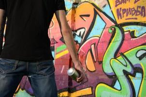 Young graffiti artist with backpack and gas mask on his neck paints colorful graffiti in pink tones on brick wall. Street art and contemporary painting process photo