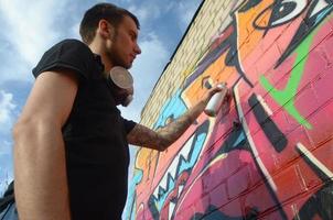 Young graffiti artist with backpack and gas mask on his neck paints colorful graffiti in pink tones on brick wall. Street art and contemporary painting process photo