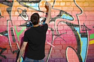 Young graffiti artist with backpack and gas mask on his neck paints colorful graffiti in pink tones on brick wall. Street art and contemporary painting process photo
