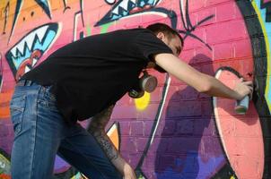 Young graffiti artist with backpack and gas mask on his neck paints colorful graffiti in pink tones on brick wall. Street art and contemporary painting process photo