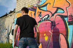 Young graffiti artist with backpack and gas mask on his neck paints colorful graffiti in pink tones on brick wall. Street art and contemporary painting process photo