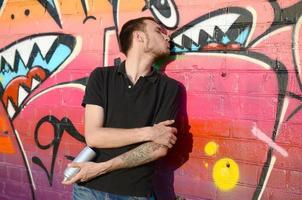 Young caucasian graffiti artist in black t-shirt with silver aerosol spray can near colorful graffiti in pink tones on brick wall. Street art and contemporary painting process photo