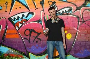 Young caucasian graffiti artist in black t-shirt with silver aerosol spray can near colorful graffiti in pink tones on brick wall. Street art and contemporary painting process photo