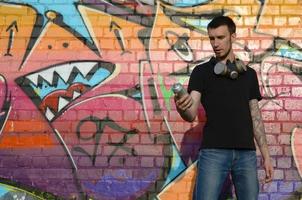 Young caucasian graffiti artist in black t-shirt with silver aerosol spray can near colorful graffiti in pink tones on brick wall. Street art and contemporary painting process photo