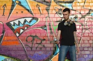 Young caucasian graffiti artist in black t-shirt with silver aerosol spray can near colorful graffiti in pink tones on brick wall. Street art and contemporary painting process photo