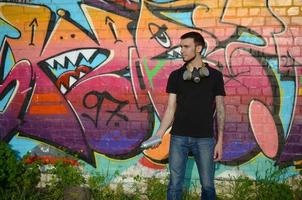 Young caucasian graffiti artist in black t-shirt with silver aerosol spray can near colorful graffiti in pink tones on brick wall. Street art and contemporary painting process photo