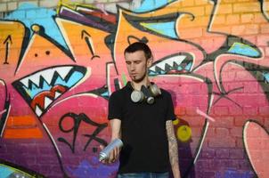 Young caucasian graffiti artist in black t-shirt with silver aerosol spray can near colorful graffiti in pink tones on brick wall. Street art and contemporary painting process photo