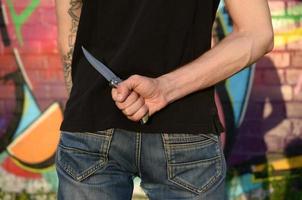 vista trasera de un joven caucásico con un cuchillo en la mano contra la pared de ladrillo del ghetto con pinturas de graffiti. concepto de fuerzas criminales y cargo de agresión foto