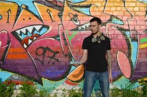 Young caucasian graffiti artist in black t-shirt with silver aerosol spray can near colorful graffiti in pink tones on brick wall. Street art and contemporary painting process photo