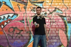 Young caucasian graffiti artist in black t-shirt with silver aerosol spray can near colorful graffiti in pink tones on brick wall. Street art and contemporary painting process photo