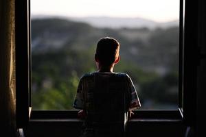 parte posterior del niño mirando desde la ventana al atardecer. foto