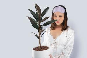 Asian woman in a white satin nightgown wearing lace robe holding rubber plant in white pot. A lady holding an air purifying plants in her hand against a gray background and space left side. photo