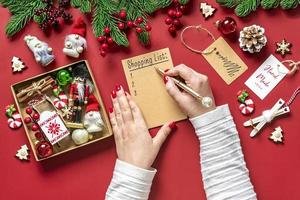 la mano femenina sostiene el bolígrafo y escribe la lista de compras navideñas, ideas de regalos en el bloc de notas blanco sobre fondo rojo vista superior plana tarjeta de vacaciones mujer haciendo lista de regalos de navidad, planificación, concepto de compra foto