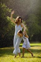 Cute little girl having fun under irrigation sprinkler with her mother photo