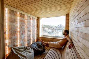 joven relajándose en la sauna y viendo el bosque de invierno a través de la ventana foto