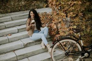 Young woman with mobile phone drink coffee to go at the stairs by the bicycle on autumn day photo