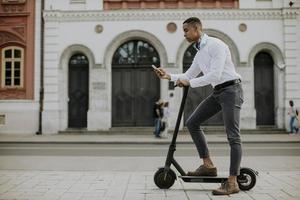 Joven afroamericano mediante teléfono móvil mientras está de pie con scooter eléctrico en una calle foto