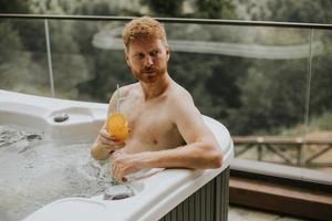 Young man enjoying in outdoor hot tub and drink fresh orange juice on vacation photo