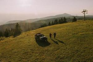 Aerial view at couple walking with backpacks by terrain vehicle over green hills photo