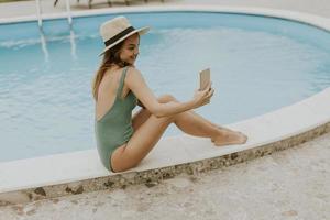 Young woman sitting by the swimming pool and taking selfie photo with  mobile phone in the house backyard