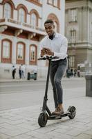 Joven afroamericano mediante teléfono móvil mientras está de pie con scooter eléctrico en una calle foto