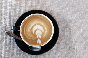 Black cup of art latte on a cappuccino coffee on beige background tsanding on a table in a cafe. Foam flower on top of the cup.Little spoon and plate. photo