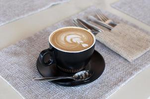 Black cup of art latte on a cappuccino coffee on beige background tsanding on a table in a cafe. Foam flower on top of the cup.Little spoon and plate. photo