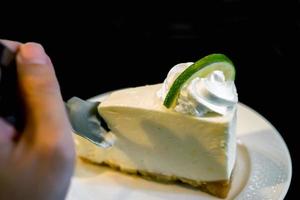 la imagen de la mano que sostiene una cucharadita está dividiendo un pastel de un pastel de crema de lima en un plato de cerámica blanca y fondo negro. foto