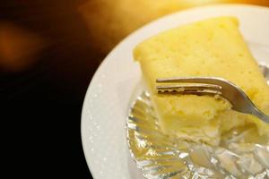 Image of fork is dividing a cake from a white chocolate cheese cake with sun flare on kitchen foil, white ceramic plate and black background. photo