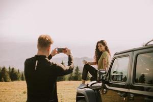pareja joven relajándose en el capó de un vehículo de terreno y tomando fotos con el teléfono móvil en el campo
