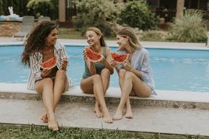 Young woman sitting on by the swimming pool and eating watermellon in the house backyard photo