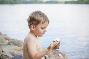 A cute blond boy appetizingly eats ice cream in the summer, sitting on the bank of the river. Cool off by the water. Funny facial expression. photo