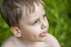 un lindo niño rubio come apetitosamente maíz en el verano, sentado en la orilla del río sobre la jugosa hierba. expresión facial divertida. foto